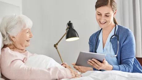 Foto einer weiblichen Pflegefachkraft, die gerade einer im Bett liegenden Senioren etwas an einem Tablet-Gerät zeigt und erklärt